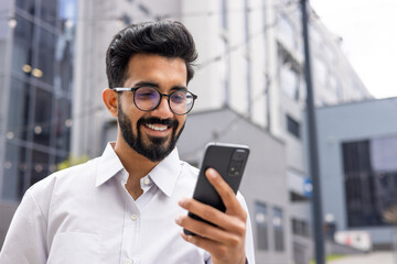 Young successful businessman walking down the street holding a phone in his hands, an Indian man using an application on a smartphone smiling contentedly from outside an office building.