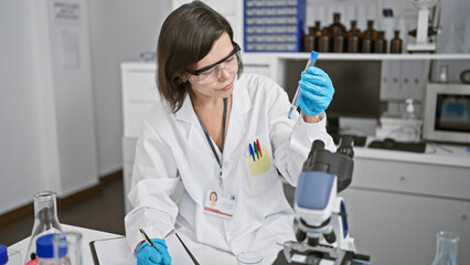 In her lab, a young, beautiful hispanic woman scientist works meticulously, jotting notes,...