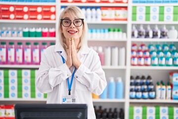 Young caucasian woman working at pharmacy drugstore praying with hands together asking for forgiveness smiling confident.