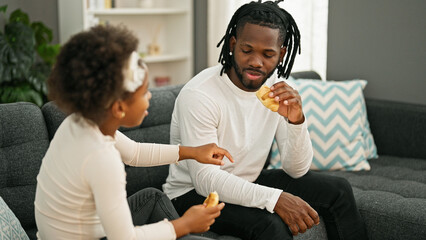 African american father and daughter sitting on sofa eating croissant smiling at home - obrazy, fototapety, plakaty