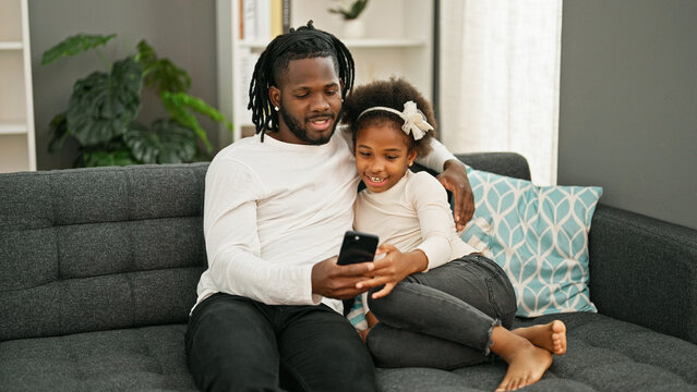 African american father and daughter using smartphone sitting on sofa smiling at home - Powered by Adobe