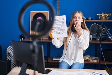 Young caucasian woman doing online music tutorial showing music sheet covering mouth with hand,...