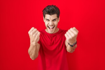 Young hispanic man standing over red background angry and mad raising fists frustrated and furious...