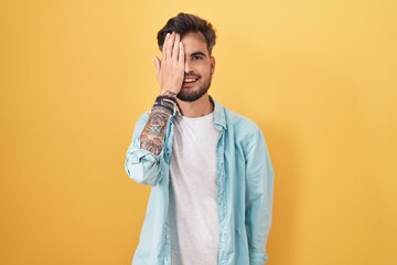 Young hispanic man with tattoos standing over yellow background covering one eye with hand, confident smile on face and surprise emotion.