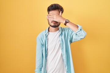 Young hispanic man with tattoos standing over yellow background covering eyes with hand, looking...