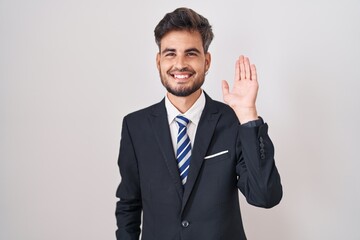 Young hispanic man with tattoos wearing business suit and tie waiving saying hello happy and smiling, friendly welcome gesture