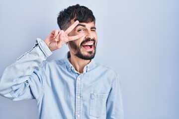 Obraz premium Young hispanic man with beard standing over blue background doing peace symbol with fingers over face, smiling cheerful showing victory