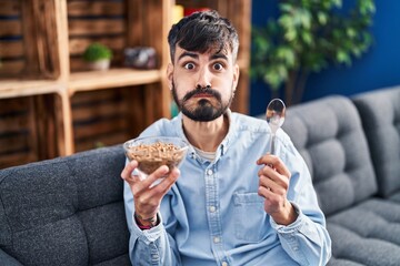 Young hispanic man with beard eating healthy whole grain cereals puffing cheeks with funny face....