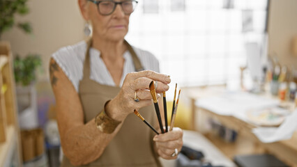 Dedicated senior grey-haired woman artist, holding paintbrushes, deeply engrossed in painting at...