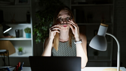 Young blonde woman business worker using laptop doing funny gesture with pen on mouth at the office