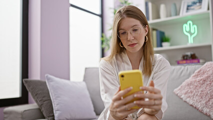 Young blonde woman using smartphone sitting on sofa with serious face at home