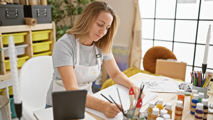 Radiant, young blonde artist joyfully sketching away on her touchpad, pouring her soul into the...