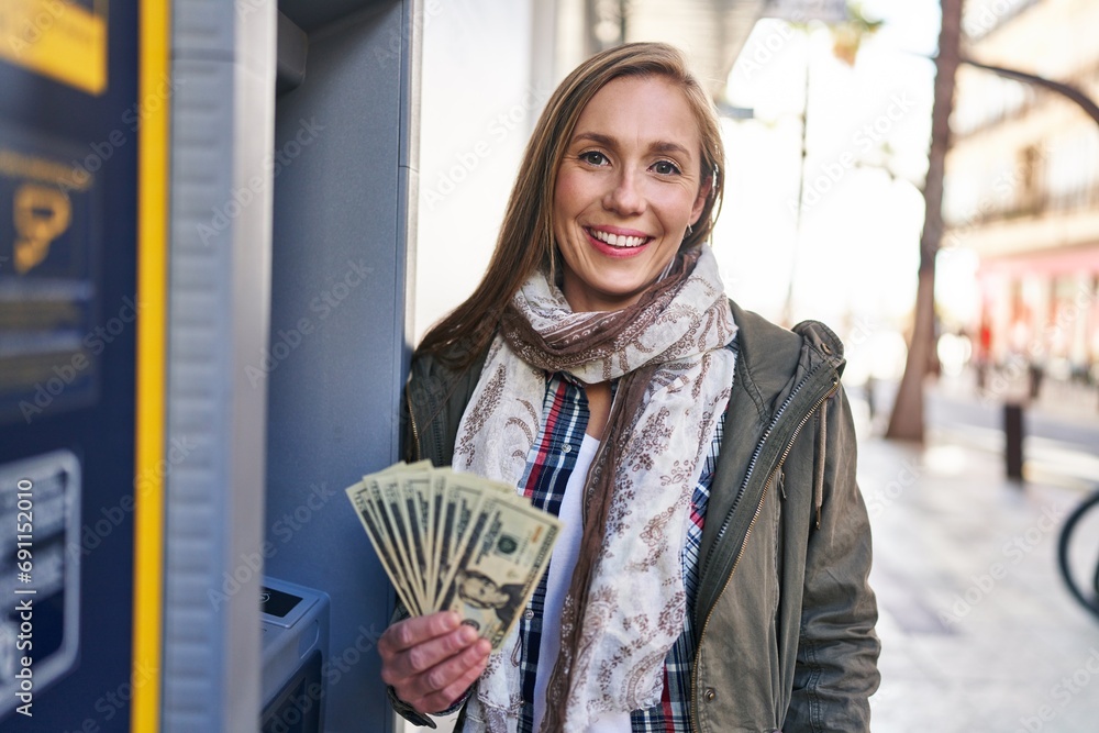 Poster Young blonde woman holding dollars banknotes from atm machine looking positive and happy standing and smiling with a confident smile showing teeth