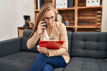 Young blonde woman psychologist talking on smartphone reading document at psychology clinic