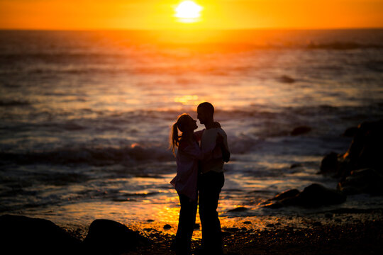 Silhouettes of a young couple in a cuddle position on the beach during a golden sunset.