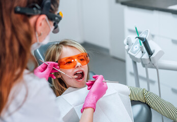 Portrait of little girl in eyeglasses sitting in stomatology chair while female dentist's palms in...