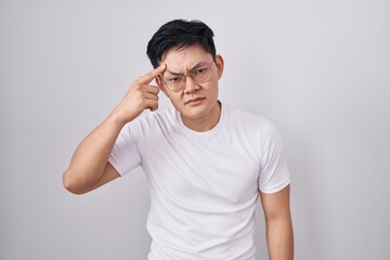Young asian man standing over white background pointing unhappy to pimple on forehead, ugly...