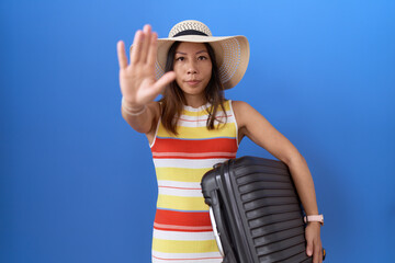 Middle age chinese woman holding suitcase going on summer vacation with open hand doing stop sign with serious and confident expression, defense gesture