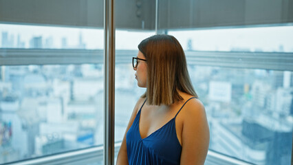 Elegant hispanic woman in glasses, admiring the city skyline from her skyscraper office, looking...