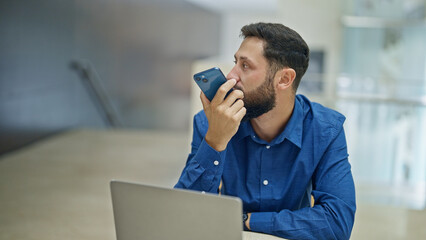 Young hispanic man business worker using laptop sending voice message by smartphone at the office