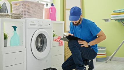 Young hispanic man technician repairing washing machine writing report at laundry room