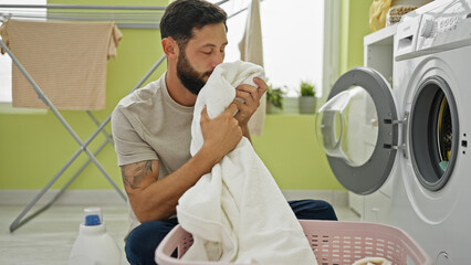 Young hispanic man washing clothes smelling clean towel at laundry room