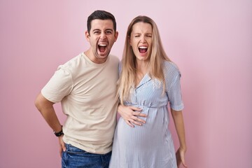 Young couple expecting a baby standing over pink background angry and mad screaming frustrated and...