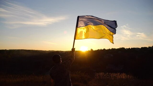 Young man in military uniform waving flag of Ukraine against sunset at background. Male ukrainian army soldier lifted national banner at countryside. Victory against russian aggression. End of war