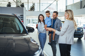 Young happy family enjoying while buying a new car in a showroom. Saleswoman at car dealership center helping family to choose new family vehicle. Family in a car dealership - obrazy, fototapety, plakaty