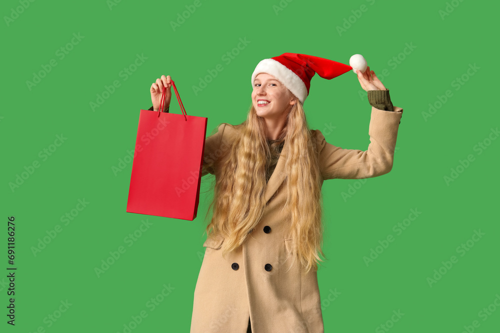 Wall mural beautiful young happy woman in warm winter clothes and santa hat with shopping bag on green backgrou