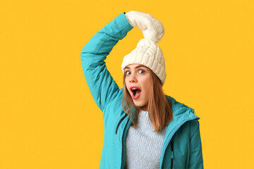 Beautiful young shocked woman in warm winter clothes on yellow background