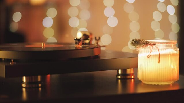 A vinyl record spins in the modern gramophone music player and plays an old disco. Close-up shot of custom vinyl turntable player with carbon tonearm. Candle and garlands in the background