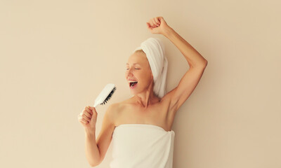 Happy cheerful middle-aged woman having fun sings with hair comb and laughing after shower drying...