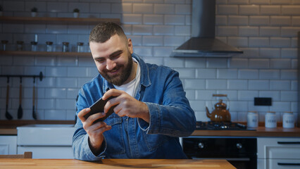 Smiling young adult man sitting against the kitchen counter playing video game on a smartphone....