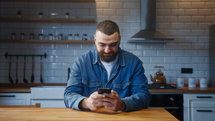 Bearded young adult man sitting against the kitchen counter typing text message by smartphone. Male...