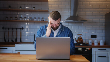 Bearded young adult man sitting against the kitchen counter having headache while using laptop....