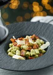 Homemade spinach pasta with fried slices of ham and parmesan cheese on holiday table with glasses of wine, background with bokeh, lights garland and glitter. Merry Christmas and New Year food, closeup