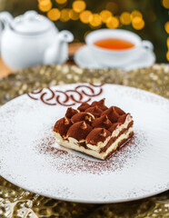 Christmas cake tiramisu sprinkled with powdered cocoa in the background a cup of tea with bokeh, lights garland and glitter. Xmas and New Year holiday