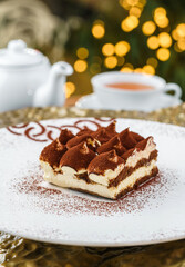 Christmas cake tiramisu sprinkled with powdered cocoa in the background a cup of tea with bokeh, lights garland and glitter. Xmas and New Year holiday, close up