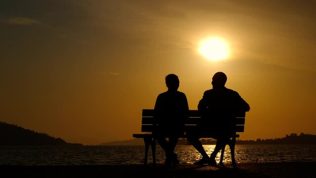 Couple silhouette resting against dusk. A view of silhouettes of man with female sitting on the bench and admire the evening sky in summer. A concept of love in family.