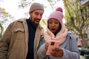 Young multiethnic couple using phone, looking at it surprised after finding out they won something...