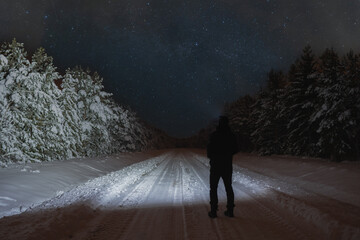 Winter night scene, a man with a flashlight in a snowy forest on the road, starry sky with clouds.