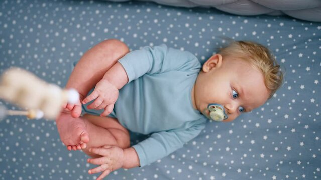 Sweet Caucasian baby boy with blond hair with pacifier in mouth. Cute infant kid lies in his crib looking at cot mobile.