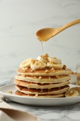 Pouring honey from spoon onto delicious pancakes with bananas and walnuts at white marble table