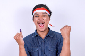 Excited Asian male wear headband, clenching fist and showing finger after voting on Indonesia...