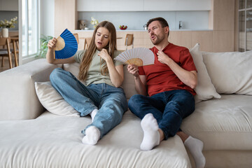 Unhappy couple sitting on sofa waving with hand fans because of broken conditioner, absence of ventilator, air blower. Wife and husband feeling unwell, dehydrated, having difficult breath, headache - obrazy, fototapety, plakaty