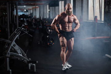 Muscular bald man posing in shorts. Bodybuilder showing off his shape in the gym. 