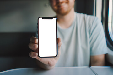 A man with a beard and mustache in a blue t-shirt is using a smartphone while traveling by Railway train, sitting in the train and looking out the window.