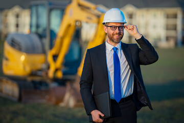 Civil engineer worker at a construction site. Mature engineer worker. Man in suit and hardhat helmet at construction site. Middle aged head civil engineer worker standing outside near excavator.