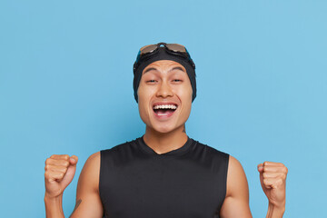 Happy man in black top and swimming cap with goggles stands on blue background with fists up, sport...